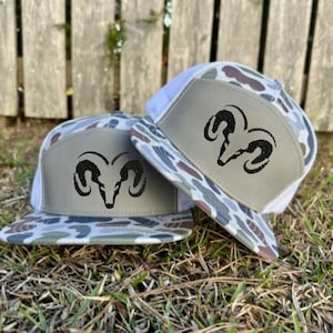 May include: Two camouflage-patterned trucker hats with white mesh backs and gray front panels. Each hat features a black ram head logo. The hats are displayed on a bed of dry grass, with a wooden fence in the background.