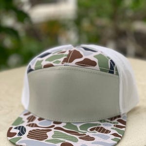 May include: A trucker hat with a camouflage pattern on the bill and top, featuring shades of brown, green, and blue. The front panel is a solid light green, and the back is white mesh. A stylish accessory for outdoor activities.