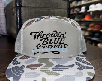 May include: A trucker hat with a camouflage pattern in shades of gray, brown, and white. The front panel is light gray with the words "Throwin' BLUE STRIPS" in black script and light blue text. The hat has a white mesh back.
