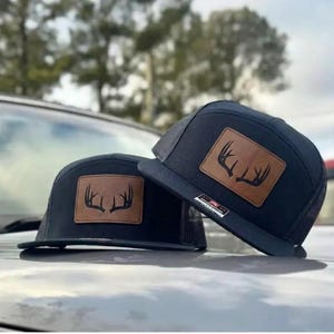 May include: Two black trucker hats with a brown leather patch featuring a deer antler design. The hats have a mesh back and a flat brim. The hats are sitting on a reflective surface.