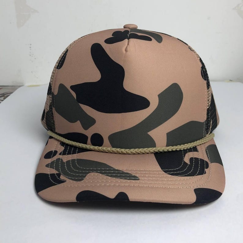 May include: A tan and black camouflage trucker hat with a curved brim and a mesh back. The hat features a rope detail along the brim and a button on top. The camouflage pattern includes shades of black, green, and tan.