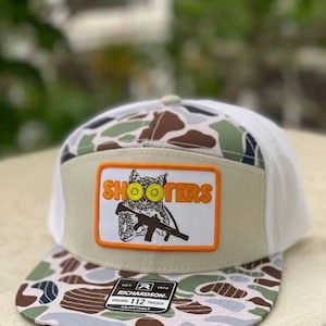 May include: A trucker hat with a camouflage pattern on the bill and a white mesh back. The front features a tan patch with the word "SHOOTERS" above an owl graphic holding a rifle. The hat is adjustable.