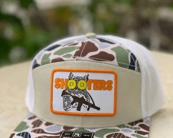 May include: A trucker hat with a camouflage pattern on the bill and a white mesh back. The front features a tan patch with the word "SHOOTERS" above an owl graphic holding a rifle. The hat is adjustable.