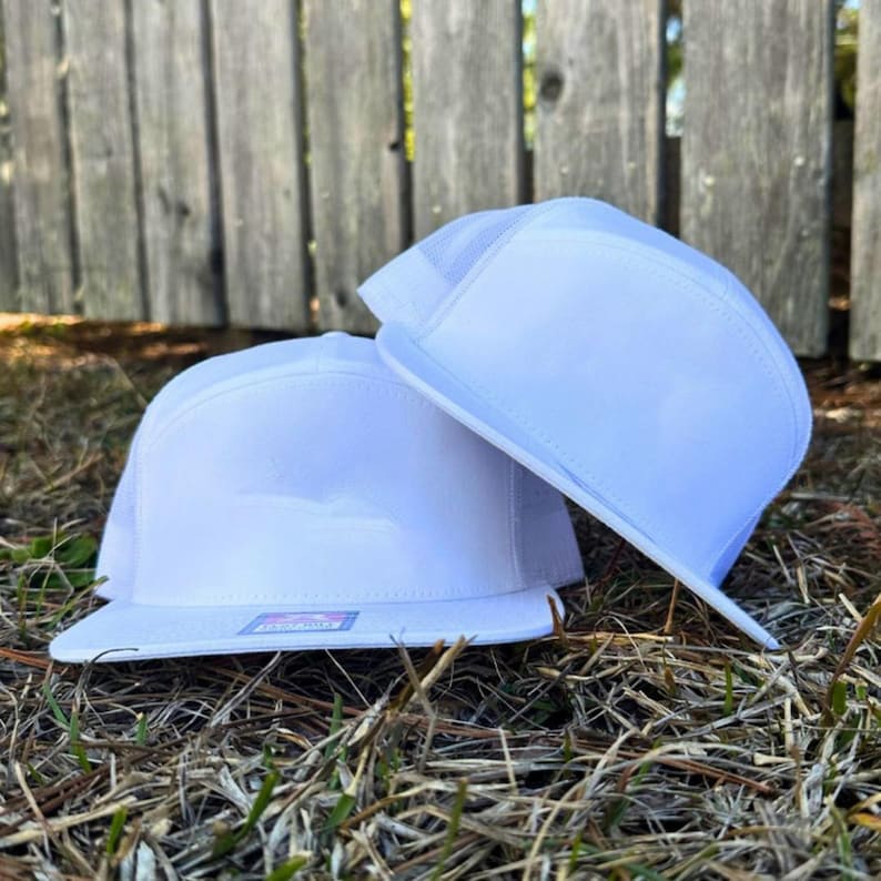 May include: Two white flat-brimmed trucker hats are stacked on top of each other. The hats have a white mesh back and a white front panel. The hats are sitting on a bed of dry grass, with a wooden fence in the background.