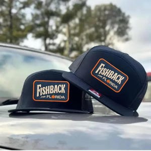 May include: Two black trucker hats with the text "Fishback for Florida" in an orange rectangle. The hats have a mesh back and a flat brim. The hats are sitting on a car.