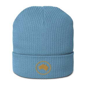 May include: A light blue beanie with a ribbed knit texture. The hat features a gold-colored embroidered emblem of a map of Australia inside a gold circle on the front. The beanie is a casual accessory.