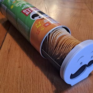 Könnte beinhalten: Eine teilweise geöffnete Pringles-Kartoffelchip-Dose mit einem individuellen weißen Deckel mit einem Cartoon-Gesicht mit Schnurrbart. Die Dose ist grün und silber. Die Chips sind im Inneren gestapelt.