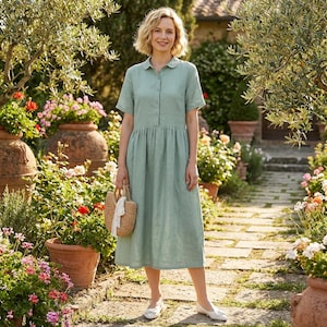 May include: A light green linen dress with short sleeves and a button-down front. The dress has a gathered waist and falls to mid-calf. The person is holding a straw handbag with a white scarf. The person is wearing white flats. The background is a garden.