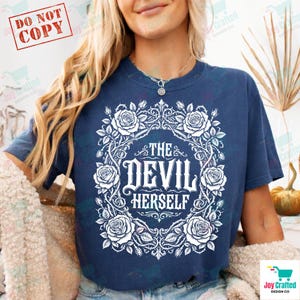 Puede incluir: Una camiseta azul marino con un diseño de corona floral blanca y el texto "THE DEVIL HERSELF" en fuente gótica. El diseño incluye rosas. La camiseta tiene mangas cortas.