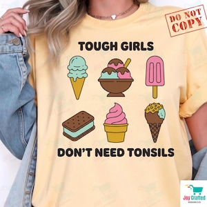 May include: A pale yellow t-shirt featuring the black text TOUGH GIRLS DON'T NEED TONSILS and a graphic of six colourful ice cream treats, including a cone, a bowl with scoops, a popsicle, an ice cream sandwich, a soft serve, and a nut-topped cone.