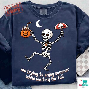 Pode incluir: Uma t-shirt azul-marinho com um gráfico de um esqueleto branco a dançar sob uma lua crescente. O esqueleto segura uma caneca de abóbora laranja e um guarda-chuva às riscas vermelhas e brancas. O texto diz, me trying to enjoy summer while waiting for fall.