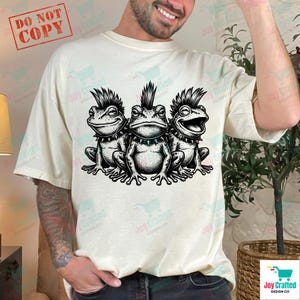 May include: Cream-colored t-shirt featuring a black and white illustration of three punk rock frogs. Each frog has a mohawk and spiked collar. The shirt has the text "Joy Crafted Design Co" printed on it.