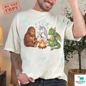 May include: Cream-colored t-shirt featuring a graphic of a Bigfoot, a unicorn, and a green dragon roasting marshmallows around a campfire. The design includes stars and a crescent moon. The words "DO NOT COPY" are in a red box in the upper left corner.