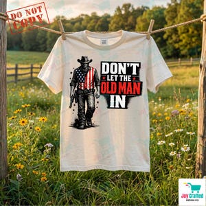 Può includere: Una maglietta color crema con la scritta "DON'T LET THE OLD MAN IN" e un'illustrazione di un cowboy con un disegno della bandiera americana sulla camicia. La maglietta è appesa a un filo per stendere i panni in un campo di fiori selvatici.