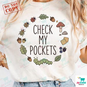 May include: Cream-coloured t-shirt with the text "CHECK MY POCKETS" in black, encircled by drawings of acorns, leaves, insects, a mushroom, and a caterpillar. The shirt has a round neckline and short sleeves.