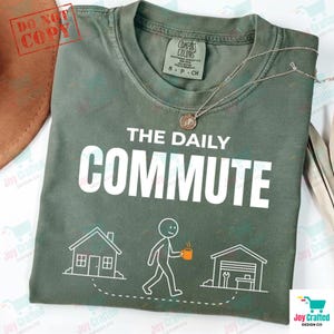 May include: A sage green t-shirt with the text "THE DAILY COMMUTE" in white. Below the text is a graphic of a stick figure walking between a house and a garage. A silver necklace with a pendant is on the shirt.