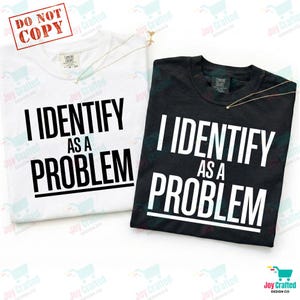 Puede incluir: Dos camisetas, una blanca y otra negra, cada una con el texto "I IDENTIFY AS A PROBLEM" en letras negras y blancas en negrita. Cada camiseta tiene un collar dorado. Las camisetas son de Joy Crafted Design Co.