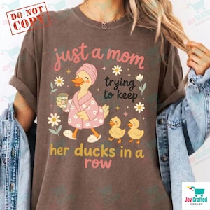 Op de afbeelding: Bruin t-shirt met de tekst "just a mom trying to keep her ducks in a row". De afbeelding toont een cartoon eend in een roze badjas en handdoek, met een koffiekopje, drie eendjes en bloemmotieven.