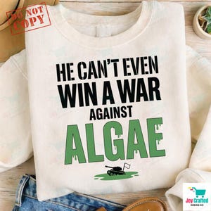 Könnte beinhalten: Ein cremefarbenes Sweatshirt mit Rundhalsausschnitt und dem schwarzen Text HE CAN'T EVEN WIN A WAR AGAINST sowie dem Wort ALGAE in großen, grünen, tropfenden Buchstaben, ergänzt durch eine kleine Grafik eines Panzers in einer grünen Pfütze.