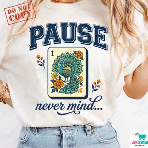 May include: A cream-colored t-shirt featuring the dark blue text PAUSE at the top and never mind... at the bottom. In the center is a rectangular tile graphic showing a peacock with blue and green feathers surrounded by orange flowers.