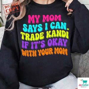 May include: A black long-sleeve sweatshirt featuring multi-colored text that reads: MY MOM SAYS I CAN TRADE KANDI IF IT'S OKAY WITH YOUR MOM. The lettering is displayed in pink, light blue, yellow, purple, and orange with a retro-style font.