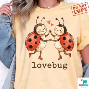 May include: A pale yellow t-shirt featuring a graphic of two cartoon ladybirds holding hands with two small red hearts above them. The text "lovebug" is printed in brown lowercase letters below the ladybirds. The shirt is worn under a denim jacket.
