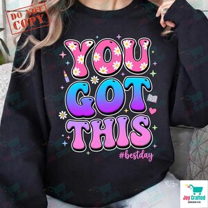 May include: Black sweatshirt with the words "YOU GOT THIS" in a colorful, retro font. The word "YOU" is pink with white daisy accents. The word "GOT" is blue and purple. The word "THIS" is pink. The hashtag "#bestday" is below.