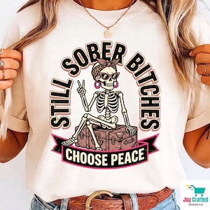 May include: A cream-colored t-shirt featuring a graphic of a skeleton wearing a leopard print headband and sunglasses, sitting on a suitcase. The skeleton makes a peace sign. Text reads: STILL SOBER BITCHES CHOOSE PEACE in black and pink lettering.