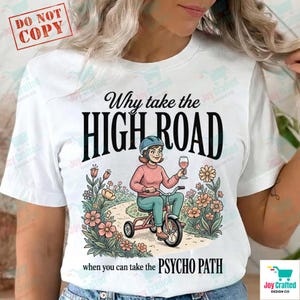 May include: White t-shirt with a graphic of a person riding a tricycle, holding a wine glass, and wearing a helmet. Text reads "Why take the HIGH ROAD when you can take the PSYCHO PATH."