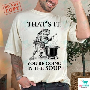 Puede incluir: Camiseta color crema con un gráfico humorístico de un chef rana. La rana, con un delantal, revuelve una olla de sopa. El texto de arriba dice "THAT'S IT." y debajo, "YOU'RE GOING IN THE SOUP." Un sello "DO NOT COPY" está en la esquina superior izquierda.