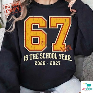 Op de afbeelding: Een zwart sweatshirt met ronde hals en een groot, verweerd geel getal 67 met een rode omlijning. Onder het getal staat de tekst 67 IS THE SCHOOL YEAR. 2026 - 2027. Het woord BRUH staat in kleine rode letters onderaan het cijfer 7.