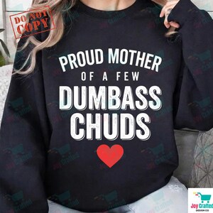 May include: Black sweatshirt with the text "PROUD MOTHER OF A FEW DUMBASS CHUDS" in white, with a red heart below the text. The words are in a bold, sans-serif font. The sweatshirt has long sleeves.