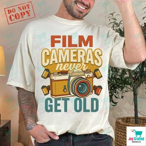 Op de afbeelding: Crèmekleurig T-shirt met een retro-ontwerp met een illustratie van een filmcamera en de tekst "FILM CAMERAS never GET OLD" in oranje en groen. Het ontwerp bevat filmrolletjes. Een "DO NOT COPY" stempel staat in de linkerbovenhoek.