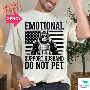 May include: Cream-colored t-shirt with a graphic featuring a gorilla wearing sunglasses and a tactical vest, set against a black and white American flag design. The text reads "EMOTIONAL SUPPORT HUSBAND DO NOT PET."