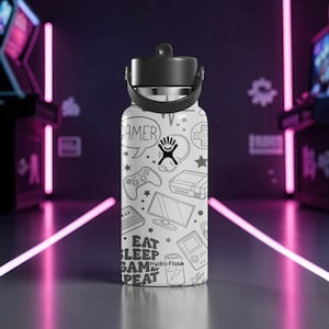Puede incluir: Botella de agua Hydro Flask blanca con tapa y asa negras. La botella está cubierta con ilustraciones en blanco y negro con temática de jugador, incluyendo un mando, estrellas y las palabras "EAT SLEEP GAME REPEAT."