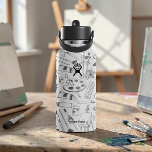 May include: A white Hydro Flask water bottle with a black lid and handle. The bottle is covered in a black and white pattern of art supplies, including paint tubes, brushes, and a palette. The Hydro Flask logo is visible.
