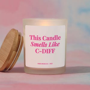 May include: A frosted glass candle with a wooden lid, the label reads "This Candle Smells Like C-DIFF" in pink text. The candle is 8 oz. and the background is a mix of pink and blue.