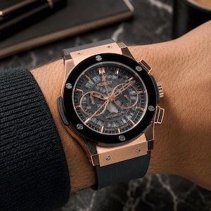 Op de afbeelding: Een automatisch Hublot-chronograafhorloge met een roségouden kast, een zwarte keramische ring vastgezet met zes H-vormige schroeven en een zwarte skeletwijzerplaat. Het horloge heeft roségouden wijzers en indexen en een zwarte rubberen horlogeband.