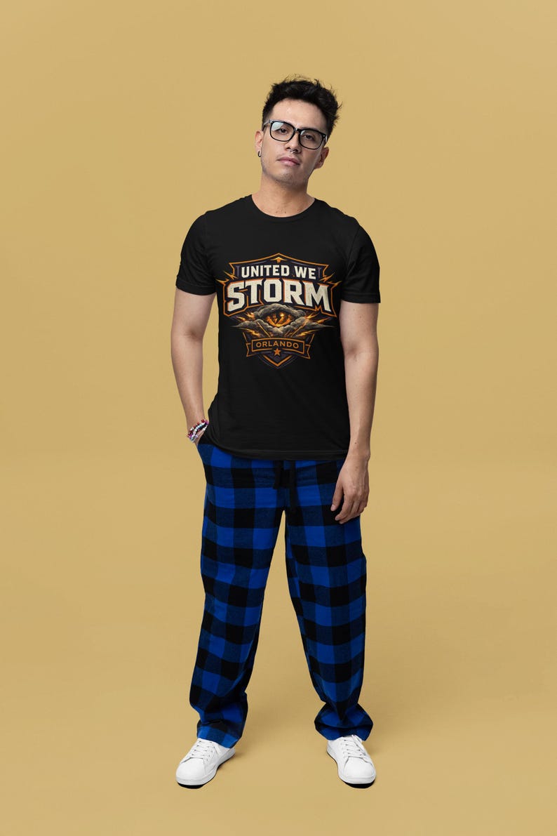 May include: Black t-shirt with the text "UNITED WE STORM ORLANDO" in orange and white. The person is wearing blue and black plaid pajama pants and white sneakers. The background is a solid tan color.
