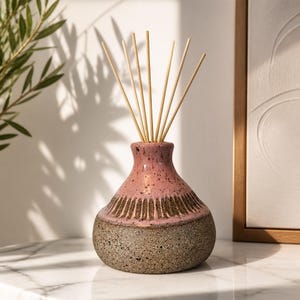 May include: A ceramic reed diffuser with a rounded base featuring a speckled gray texture on the bottom half and a smooth pink glaze on the top half. A band of dark brown vertical etched lines separates the two colors. Seven light wooden reeds.