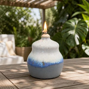 May include: A ceramic oil lamp with a rounded base and a narrow neck, featuring a gradient glaze that transitions from a matte gray bottom to a vibrant blue band and a speckled white top, topped with a lit wick emitting a small, steady flame.