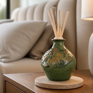 May include: A green ceramic reed diffuser bottle with a rounded body and a textured floral relief pattern on the front. The bottle features a narrow neck holding several light-colored wooden diffuser reeds extending upwards from the opening.