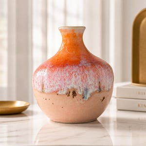 May include: A ceramic vase with a bulbous body and a narrow neck. The upper portion features a vibrant orange and white drip glaze, while the lower section displays a matte, textured peach-colored finish with visible small dark speckles.