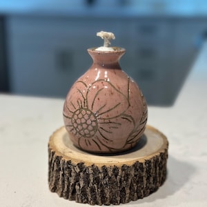 May include: A small, round, dusty pink ceramic oil lamp with a narrow neck and a white wick. The surface of the lamp features a hand-carved floral pattern with dark outlines. The lamp is resting on a circular, natural wood slice with visible bark.