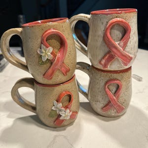 May include: Three speckled ceramic mugs with a tan base and a coral-colored interior rim. Each mug features a raised coral awareness ribbon. Two of the mugs are decorated with small white flowers with green leaves attached to the ribbon.