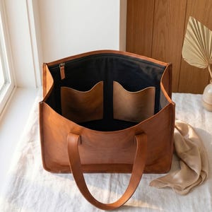Könnte beinhalten: Offene, braune Ledertasche mit schwarzem Innenfutter und zwei Taschen. Die Tasche hat einen Reißverschluss und gebogene Griffe. Sie steht auf einer weißen Oberfläche. Ein praktisches Accessoire für den Alltag.