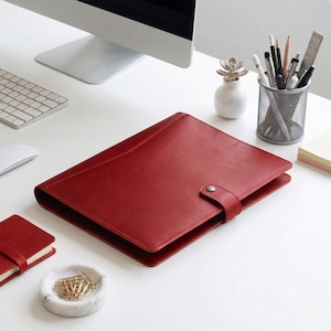 Puede incluir: Una carpeta portafolio de cuero rojo intenso con una correa de cierre de botón a presión. El organizador de documentos rectangular presenta costuras suaves a lo largo de los bordes y un diseño sencillo para organizar papeles y documentos.