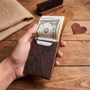 May include: A dark brown leather wallet with a textured finish, featuring a flap closure with a single metal snap. The wallet contains a one-dollar bill and two white credit cards with the text CREDIT CARD printed on the top edge of each card.