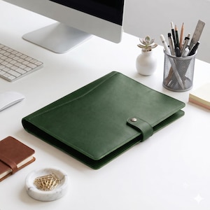 May include: A dark green leather portfolio folder featuring a smooth, matte finish and a secure strap closure with a metal snap button. The rectangular folder is positioned centrally, showcasing clean stitched edges and a minimalist design.