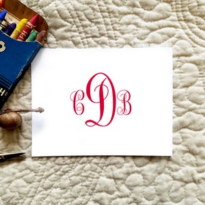May include: A white card with a red monogram featuring the letters C, D, and B in a decorative script. The card is on a textured, off-white surface, with a blue crayon box and other items in the background.